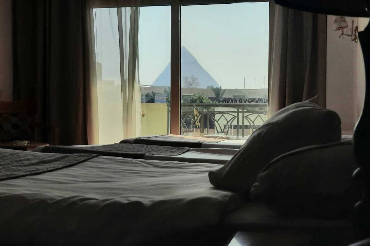 A hotel room with a bed, a window with a view of a pyramid, and a balcony with a railing.