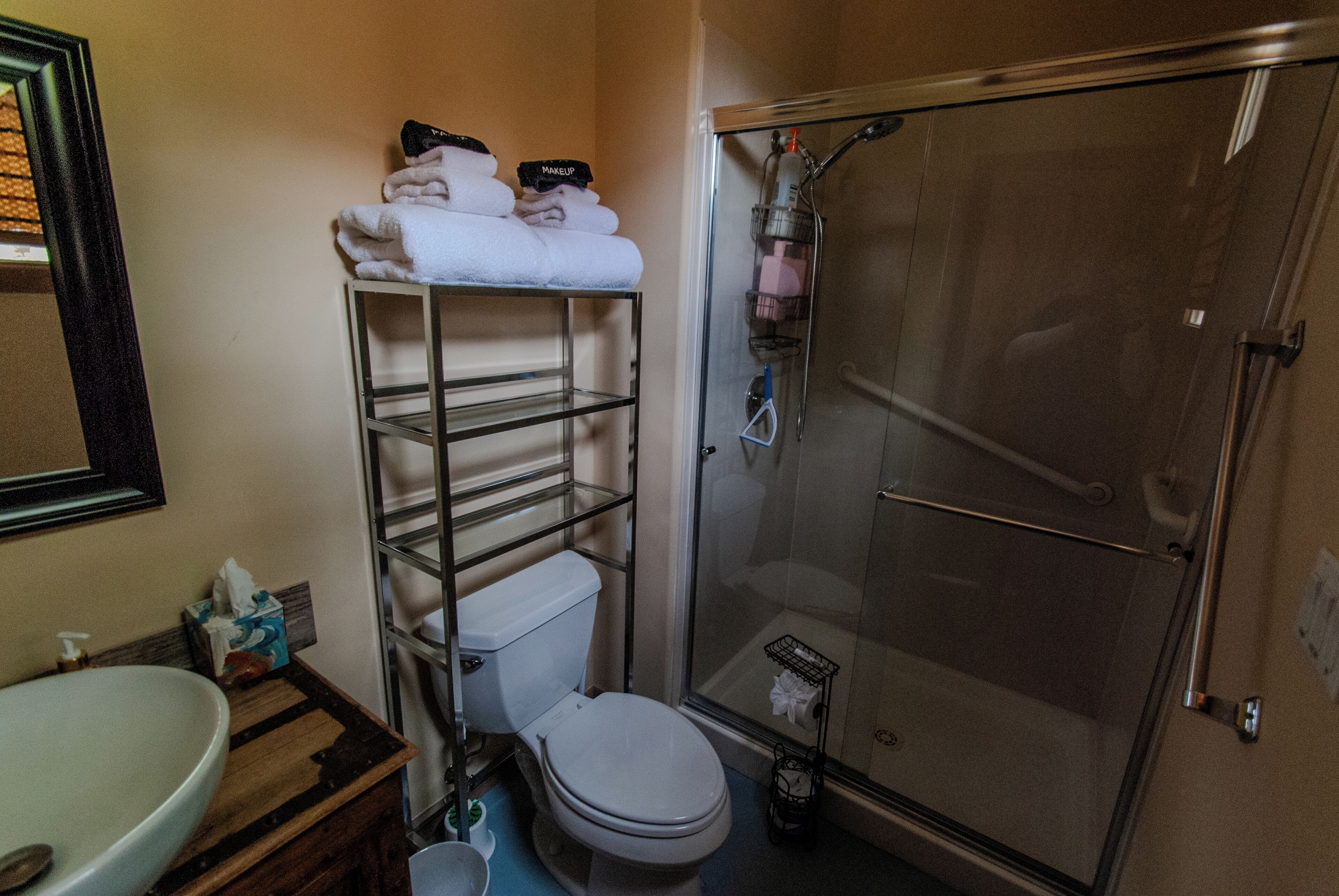 A bathroom with a toilet, sink, and shower. There is a shelf with towels and toiletries.
