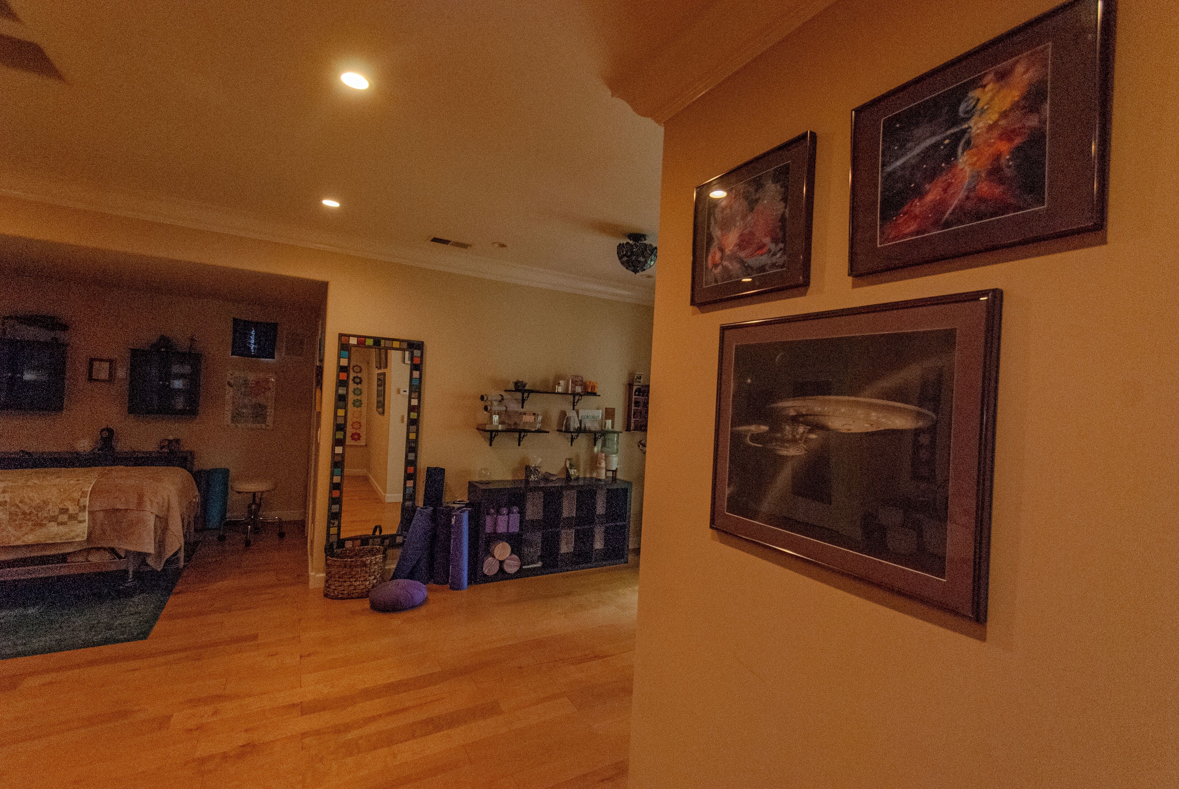 A room with wooden flooring, a bed, a desk, and a mirror. There are framed pictures on the wall and a shelf with various items.