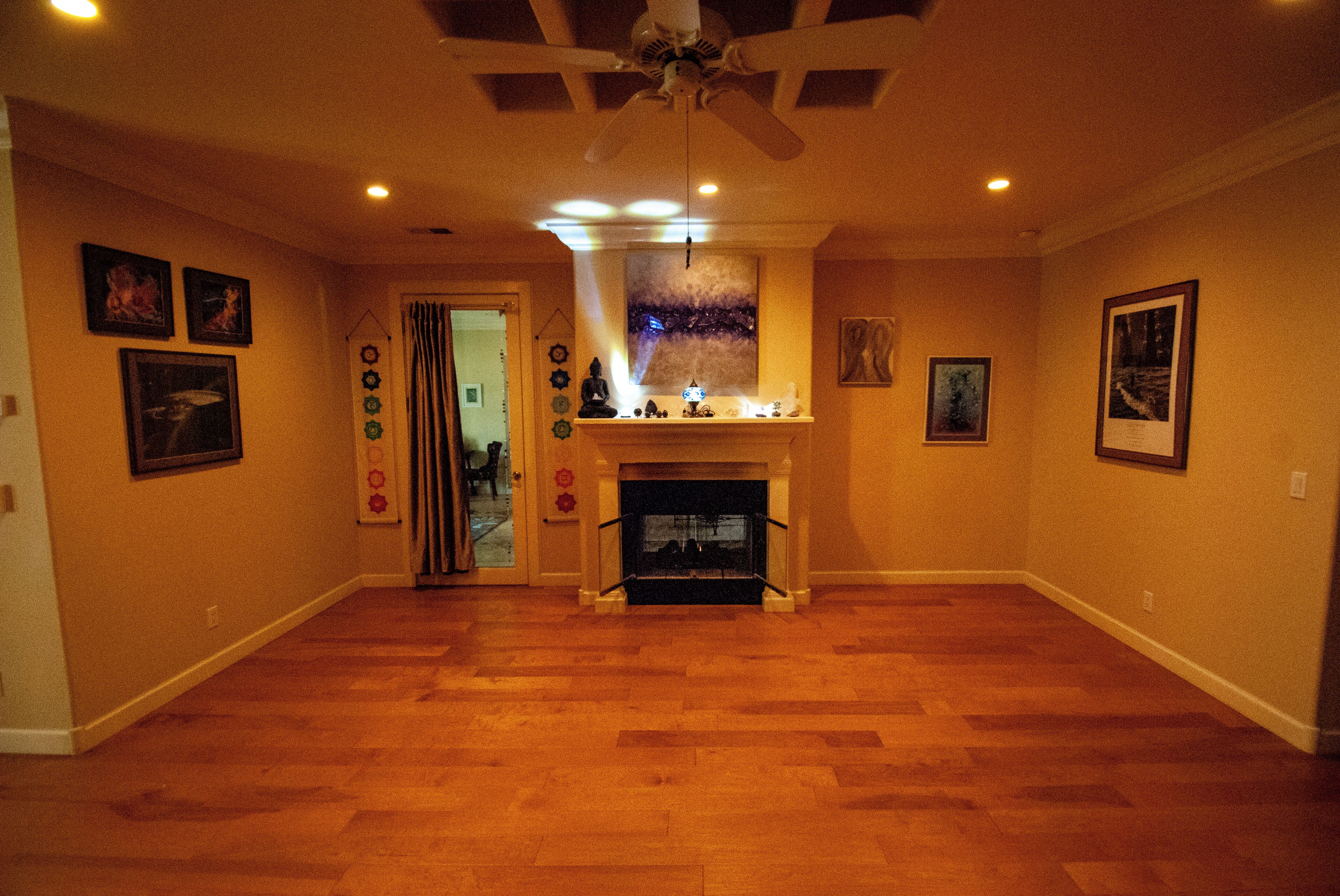 A spacious living room with hardwood flooring, a fireplace, ceiling fan, and various wall decorations.