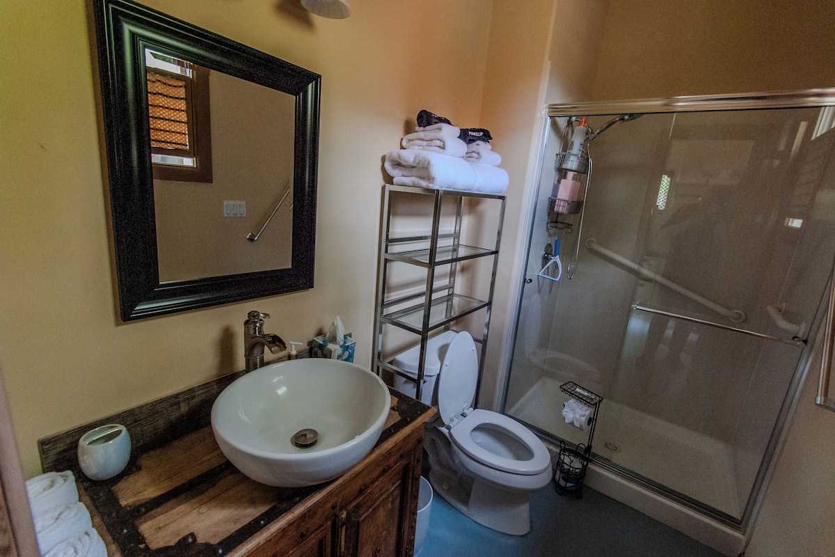 A bathroom with a wooden vanity, a large sink, a toilet, a glass shower enclosure, and a towel rack with towels.