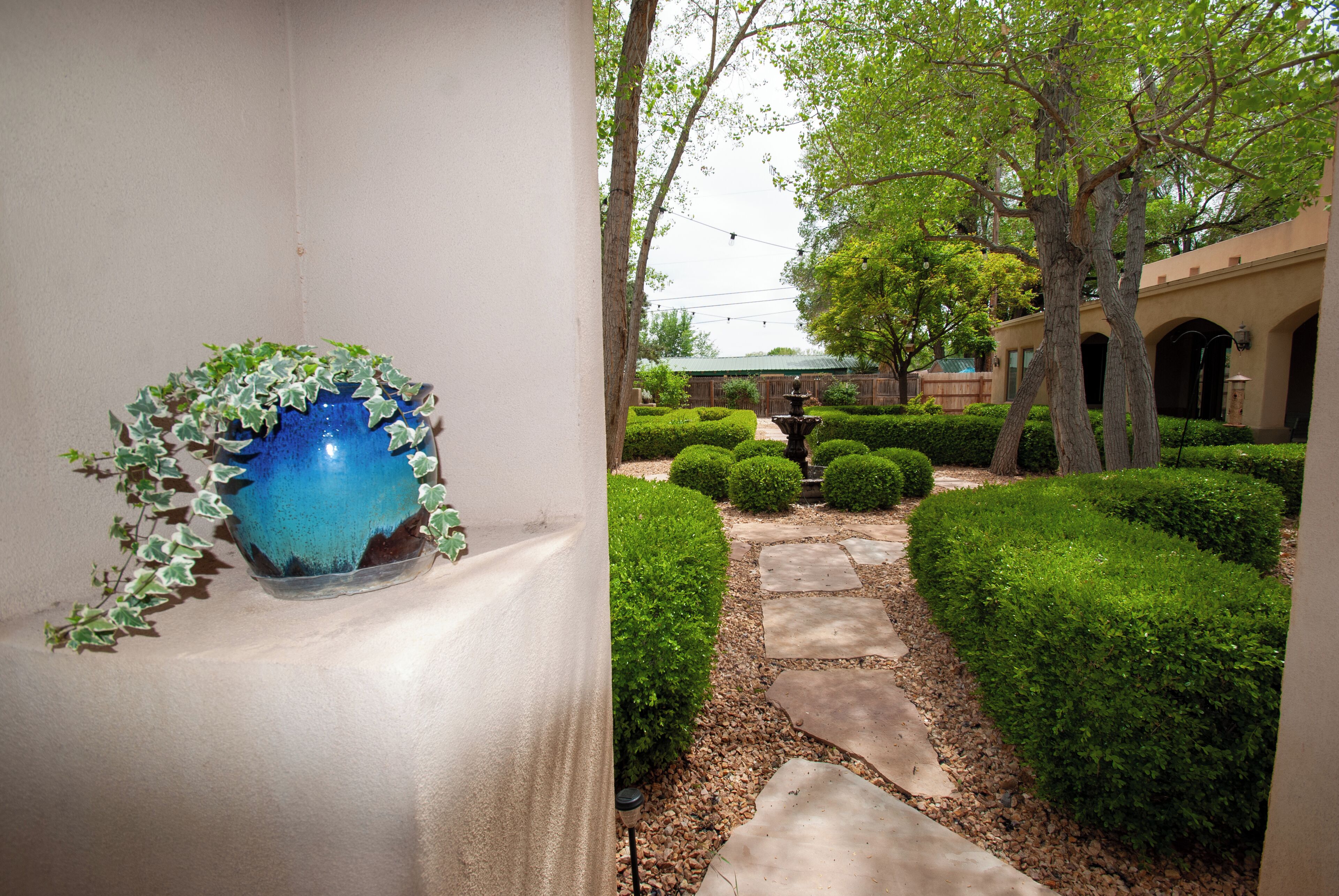 Un jardín con un camino de piedra, setos cuidados y un jarrón azul con follaje verde.