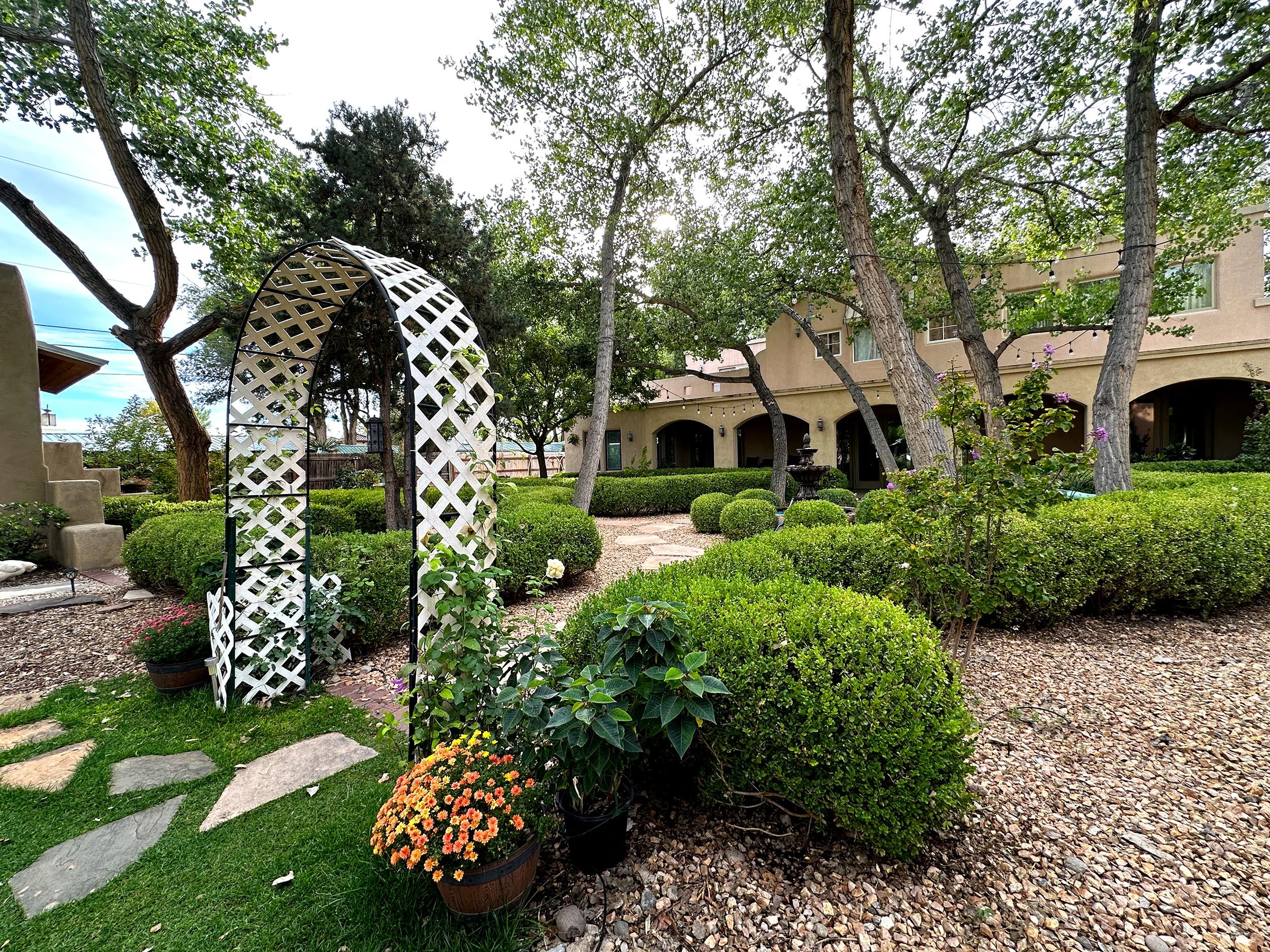 A landscaped garden with a white lattice archway, neatly trimmed hedges, and a variety of potted plants.