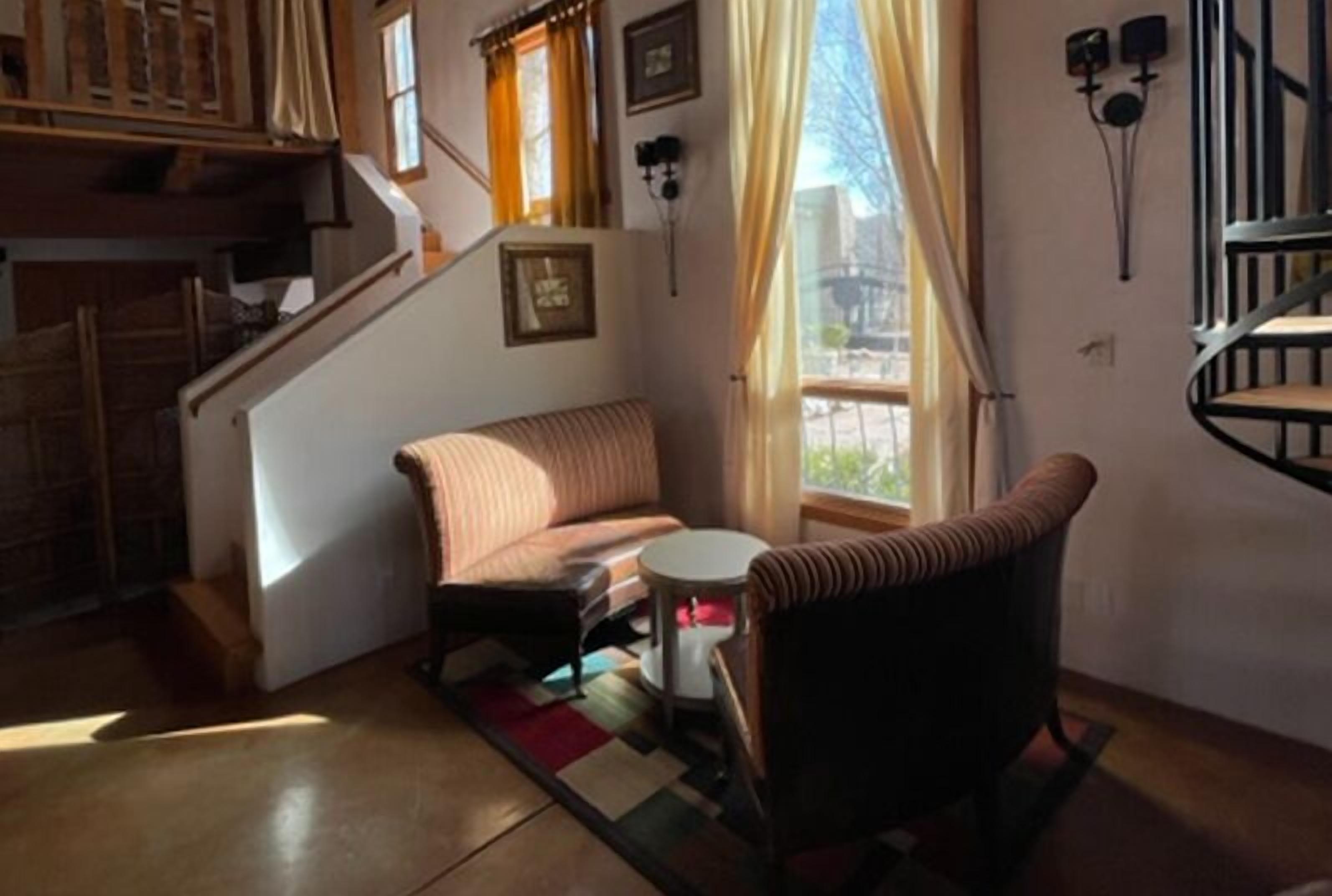 Un salon confortable avec un canapé, un fauteuil et une petite table. On aperçoit un escalier au fond.
