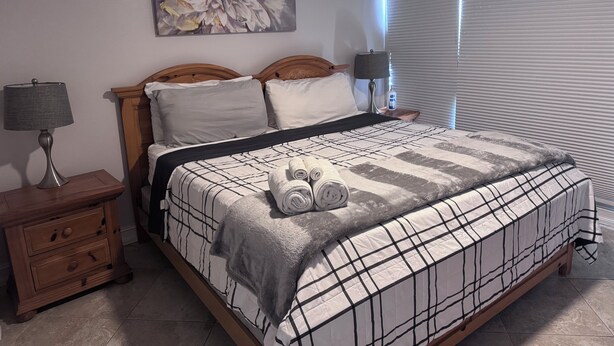 A neatly made bed with a plaid bedspread, a wooden headboard, a nightstand with a lamp, and a painting on the wall. - Great 2 BR Condo, 7 minutes to Siesta Key (Sarasota)
