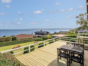 Una terraza de madera con mesa y sillas, con vistas a un cuerpo de agua y una zona residencial.