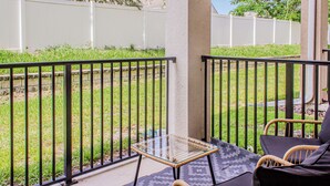 A balcony with a glass table and wicker chairs, surrounded by a white fence and greenery.