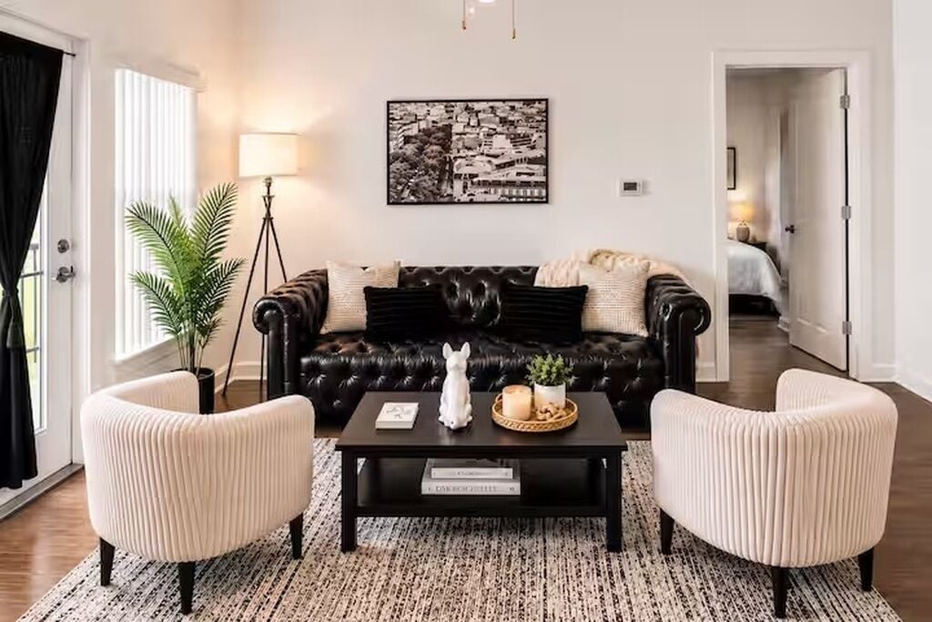 A living room with a black leather sofa, two white armchairs, a coffee table with decorative items, a floor lamp, and a framed cityscape picture on the wall.