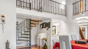 A spacious loft-style apartment with a wooden staircase, a dining area with a red table, and a kitchen visible through an open door.