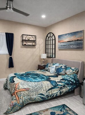 Quarto com cama, mesa de cabeceira, luminária, janela com cortinas, ventoinha no teto, prateleira na parede e um quadro.