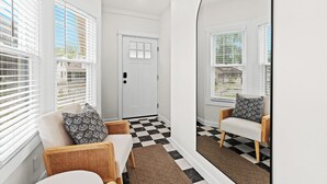 A hallway with a checkered floor, white walls, and a door. There are two wicker chairs with cushions and a mirror on the wall.