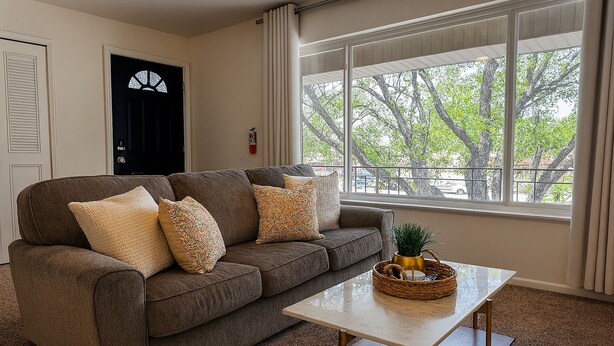 Ein Wohnzimmer mit einem braunen Sofa, mehreren Kissen, einem weißen Couchtisch und Blick auf Bäume durch große Fenster.