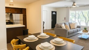 Eine moderne Küche mit Einbauofen und Kühlschrank, ein Essbereich mit dunklem Tisch und gelben Stühlen sowie ein Wohnzimmer mit grauem Sofa und Couchtisch.