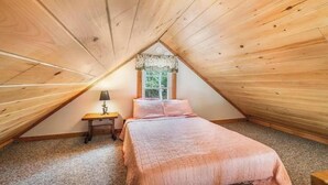Een slaapkamer met een schuine kap, een raam met kantgordijnen, een bed met een roze deken en een nachtkastje met een lamp.