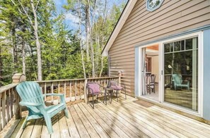 Une terrasse en bois avec des sièges d’extérieur, une porte vitrée donnant sur une pièce et un paysage boisé en arrière-plan.