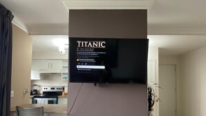 A modern living room with a flat-screen TV mounted on a wall, a dining area with a table and chairs, and a kitchen in the background.