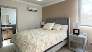 A bedroom with a bed, bedside table, nightstand, air conditioning unit, and a window.
