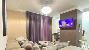 A modern living room with a beige sectional sofa, a dining area with a round table, and a flat-screen TV mounted on the wall.