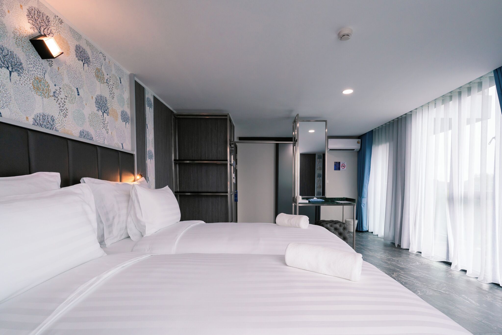 A modern hotel room with a large bed, a bedside table, a mirror, and a view of the outside through sheer curtains.