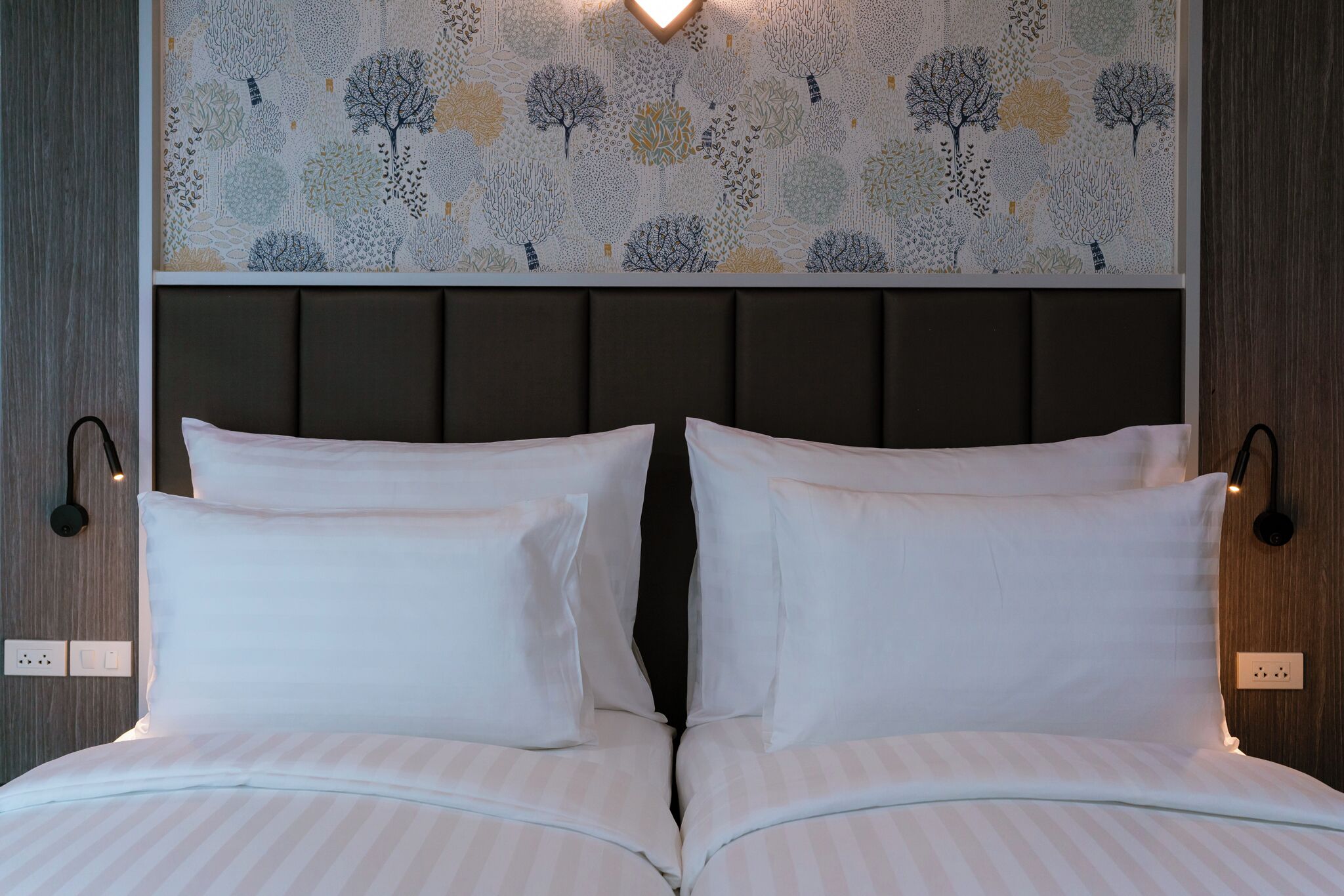 A neatly made bed with white pillows and striped bedding, a headboard with a tree-patterned wallpaper, and wall-mounted reading lights.