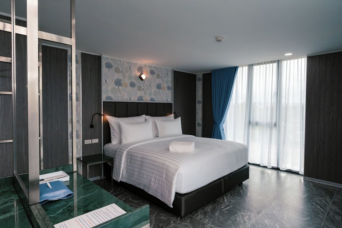 A modern hotel room with a large bed, a desk with a chair, a nightstand, and a large window with curtains.
