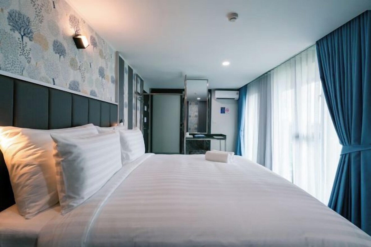 A hotel room with a large bed, a headboard with a light fixture, a desk with a chair, and a window with curtains.