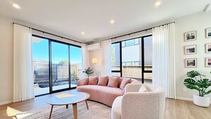 Un salon moderne avec un canapé, un fauteuil et une table basse ronde. De grandes fenêtres offrent une vue sur l’extérieur.