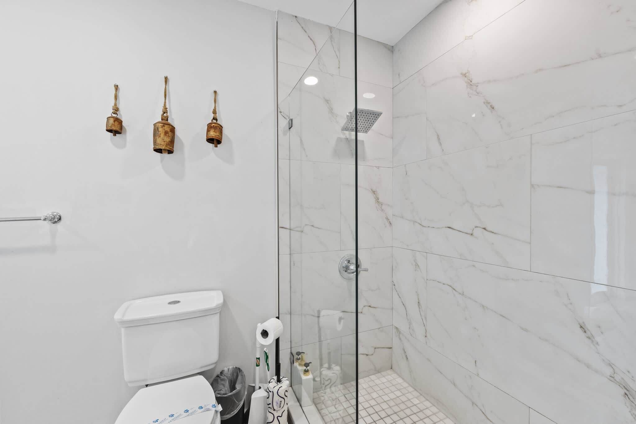 Un baño moderno con cabina de ducha de vidrio, azulejos de mármol blanco y un inodoro con dispensador de toallas de papel.