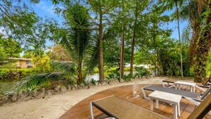 A wooden deck with lounge chairs and a small table, surrounded by lush greenery and a body of water. - Villa Venecia | Sand Terrace | Heated Pool | Waterfront (Miami)