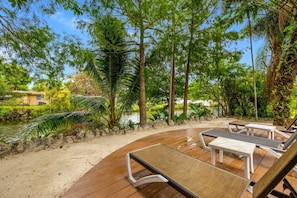A wooden deck with lounge chairs and a small table, surrounded by lush greenery and a body of water.