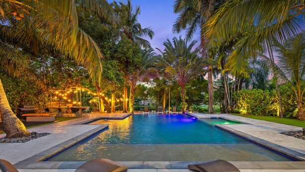 A swimming pool with lounge chairs, surrounded by palm trees and string lights. - Villa Venecia | Sand Terrace | Heated Pool | Waterfront (Miami)