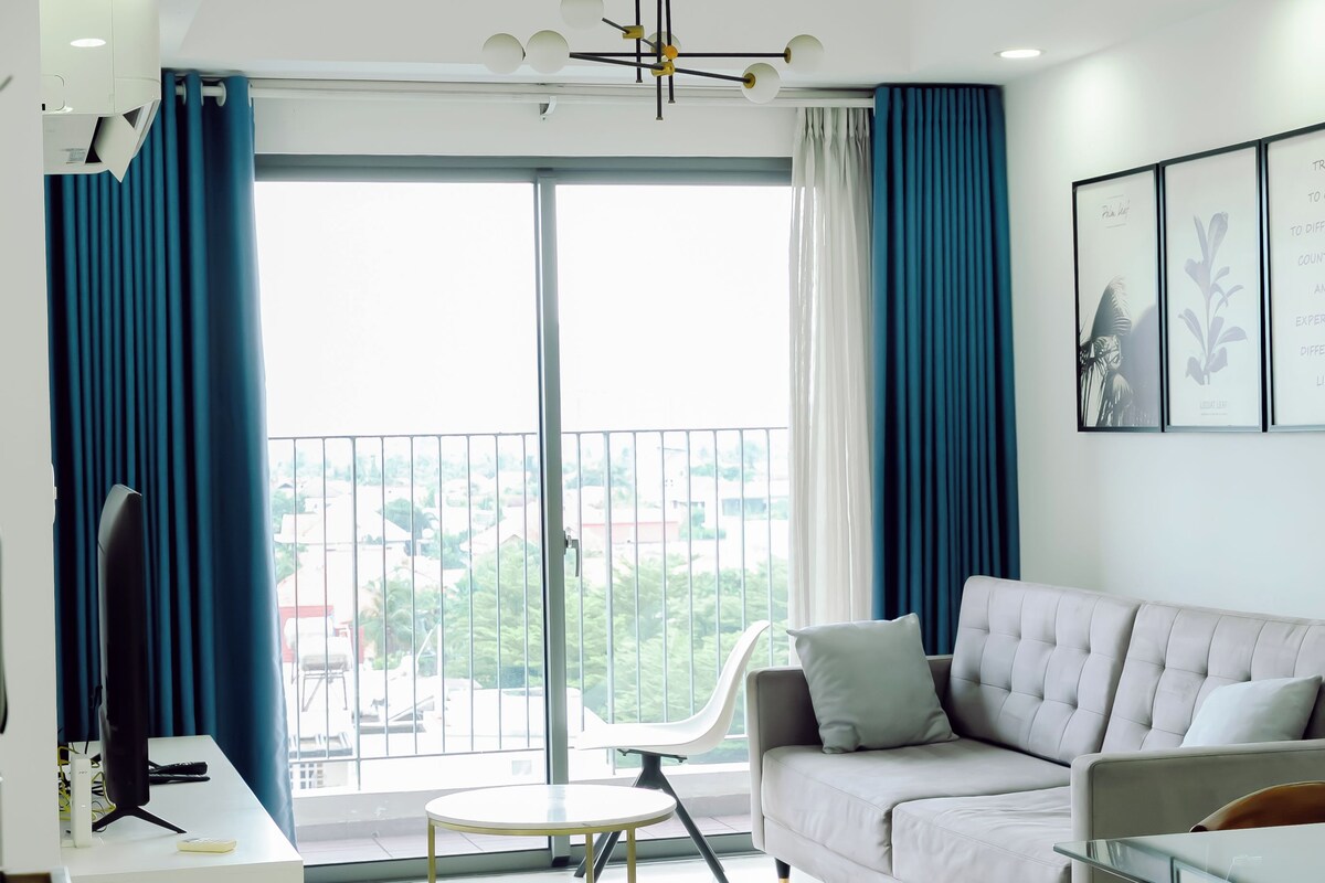 A modern living room with a sofa, a coffee table, and a TV. There is a balcony with a view of the city.