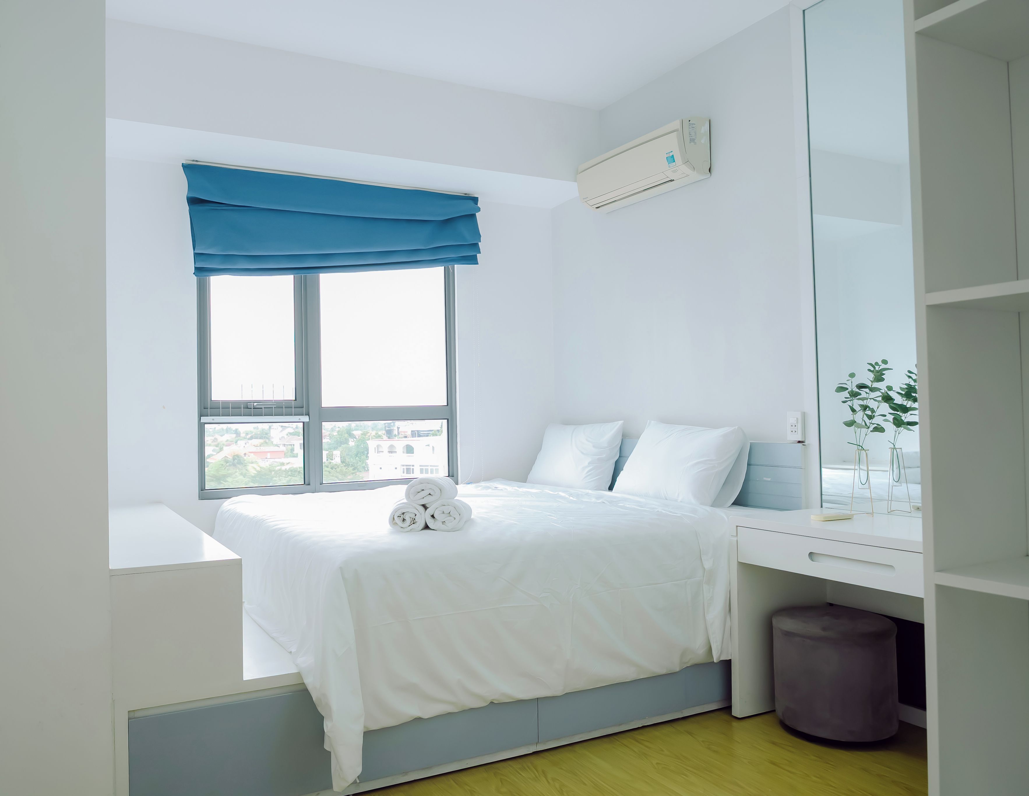 A hotel room with a bed, a desk, a window with blue curtains, and a view of buildings outside.