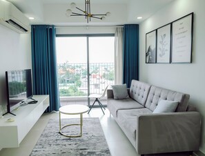 A modern living room with a flat-screen TV, a sofa, a small round table, and a view of the city through large windows. - Masteri Thao Dien Apartment Complex (Ho Chi Minh City)