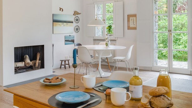 Een lichte, open woon- en eetkamer met een houten ontbijttafel, een open haard en grote ramen met uitzicht op groen.