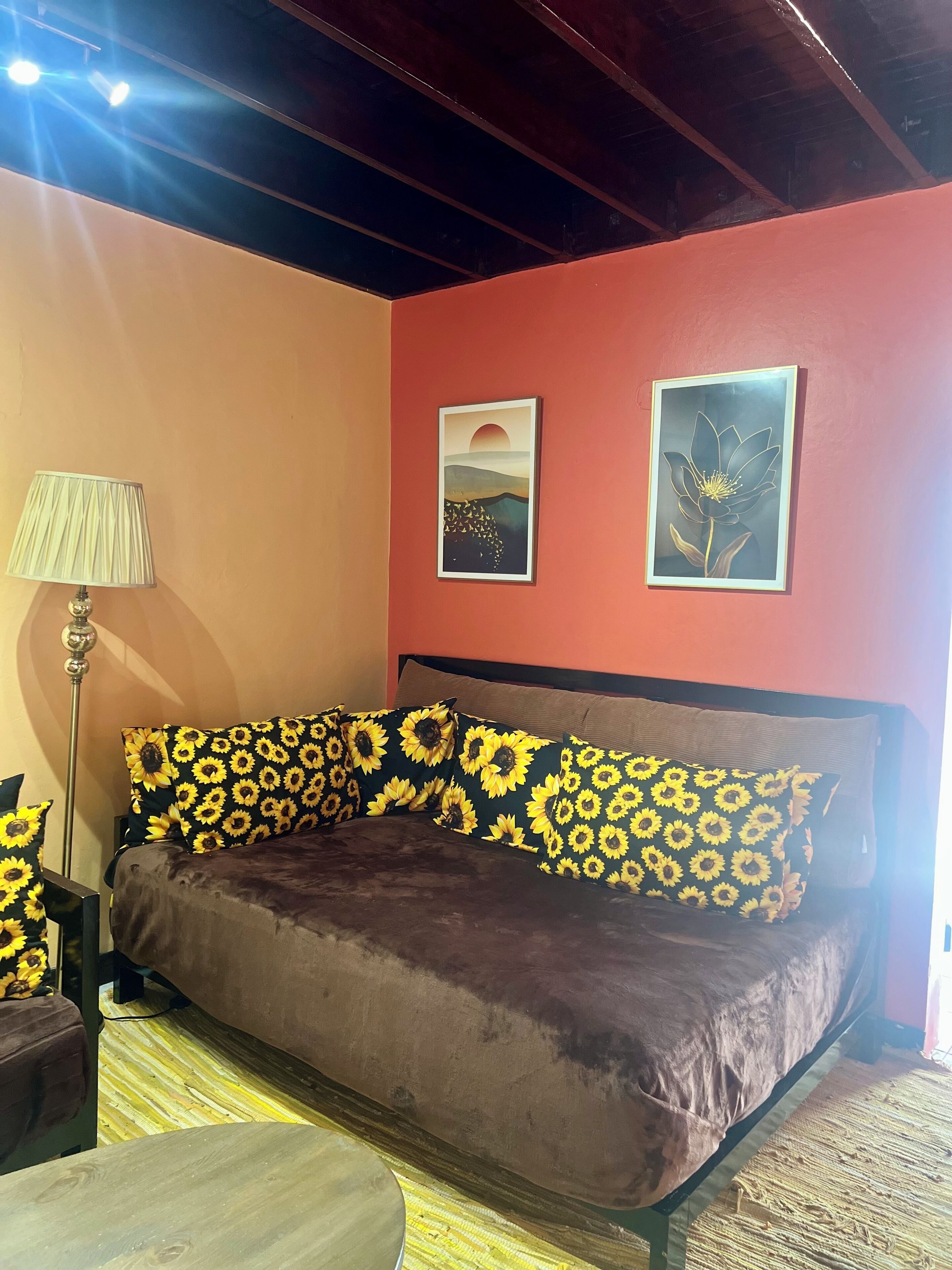 Ein Zimmer mit einem braunen Sofa, Kissen mit Sonnenblumenmuster, einer Lampe und zwei gerahmten Bildern an der Wand.