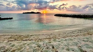 A beach with clear water, a sandy shore, and a sunset in the background.
