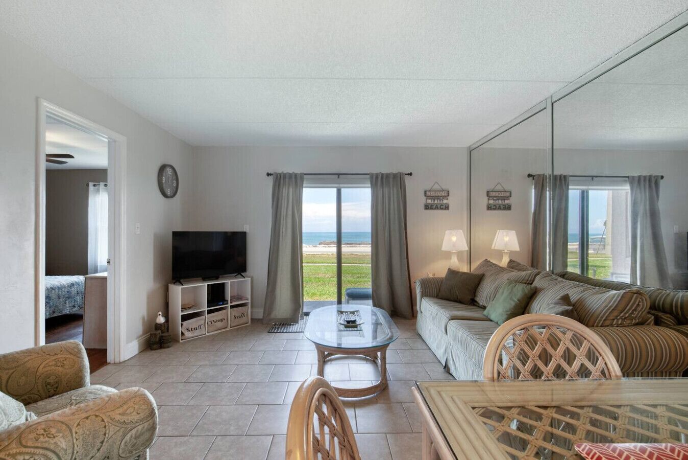 A living room with a TV, sofa, and dining table. There is a view of the beach through the windows.