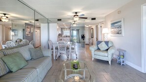 A spacious living area with a sofa, a glass coffee table, and a dining table with chairs. There is a large mirror reflecting the room, and a ceiling fan is visible. - Beach, Don’t Kill My Vibe (Ormond Beach)