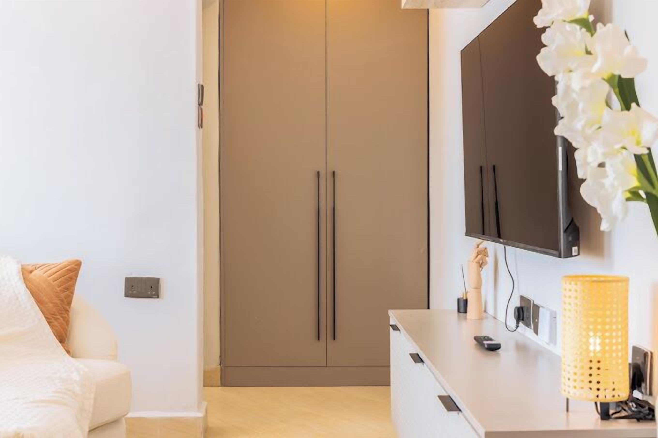 Un salon moderne avec un canapé beige, un téléviseur à écran plat fixé au mur, une petite table avec une lampe et une armoire aux portes coulissantes.
