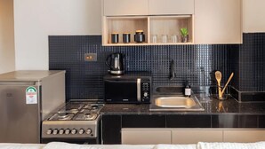 A compact kitchen with a gas stove, a microwave, a coffee maker, and a sink. There is a shelf above the sink with cups and a plant. - Lux Suites Studio City Apartments (Naivasha)