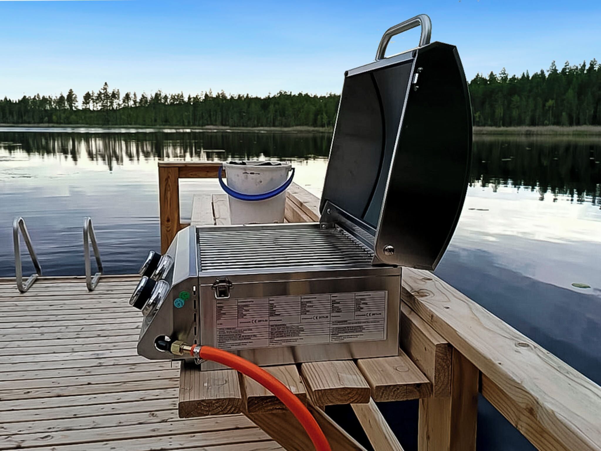 A portable grill on a wooden dock by a calm lake with a clear sky.