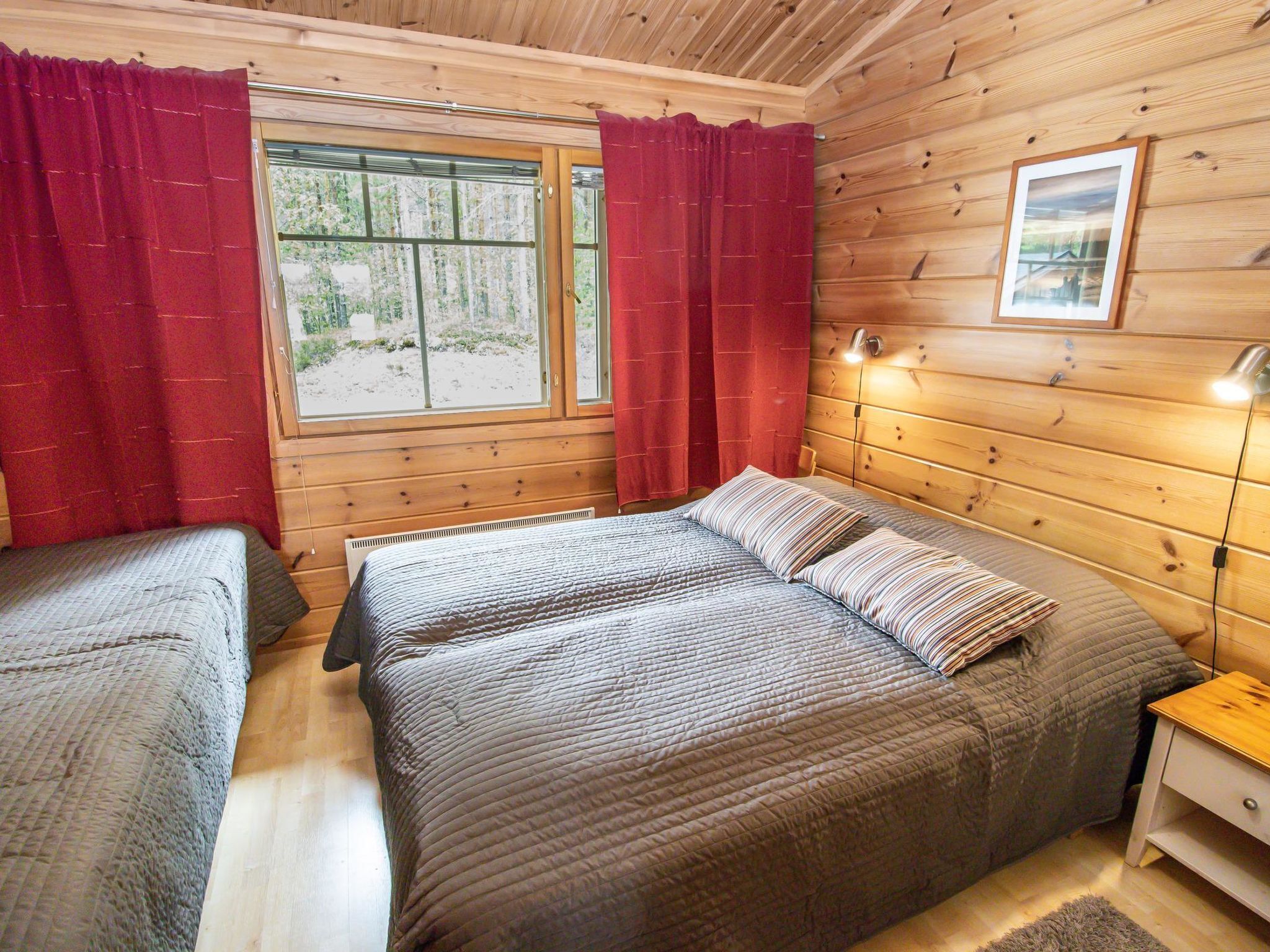 Une chambre dans un chalet en bois, avec deux lits, une fenêtre avec des rideaux, une table de chevet et un tableau encadré au mur.