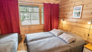 A wooden cabin bedroom with two beds, a window with curtains, a bedside table, and a framed picture on the wall.