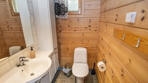 A wooden-paneled bathroom with a white sink, mirror, toilet, and a small window with a floral curtain.