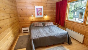 Un dormitorio de cabaña de madera con una cama, una mesita de noche, un cuadro enmarcado, una ventana con cortinas rojas y vista a otra cabaña.