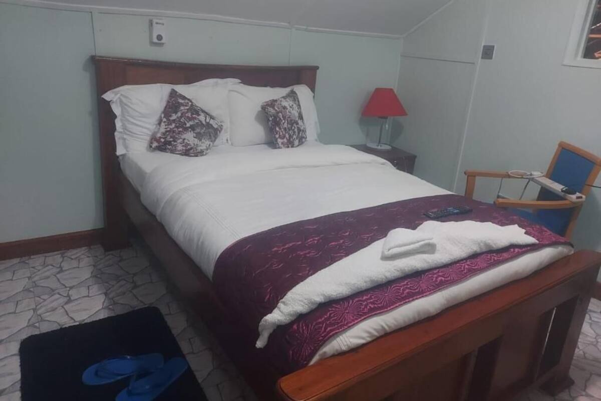A neatly made bed with a white and purple bedspread, a wooden headboard, and two pillows. There is a red lamp on a bedside table, a chair, and a small table with a red tablecloth.