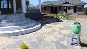A stone staircase leading to a covered porch, a green barrel on a stool, and a house with a stone facade and a tiled roof. - Kimberly Guest House Naivasha (Naivasha)