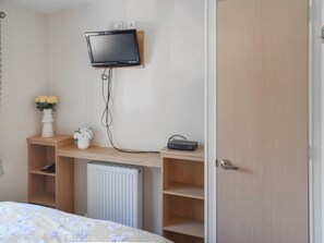 Un dormitorio con cama, televisor montado, radiador y estante con un florero y un control remoto.