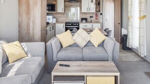 Un espai habitable reduït amb un sofà gris, una taula baixa de fusta, i una cuineta al fons.